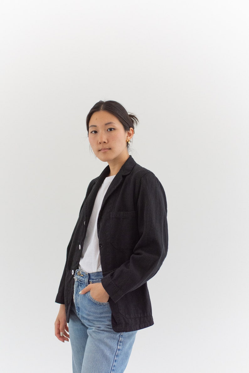 Op de afbeelding: Een vrouw draagt een zwart blazer over een wit t-shirt en een blauwe jeans. Ze staat voor een witte achtergrond.