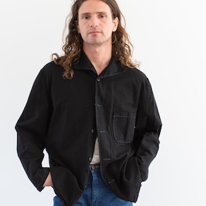 Puede incluir: Una camisa negra de manga larga con cierre de botones y un solo bolsillo en el pecho izquierdo. La camisa está hecha de una tela suave y texturizada.