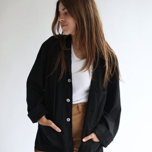 May include: A black long-sleeved button-up shirt with a relaxed fit and pockets. The shirt is worn over a white t-shirt and brown trousers.