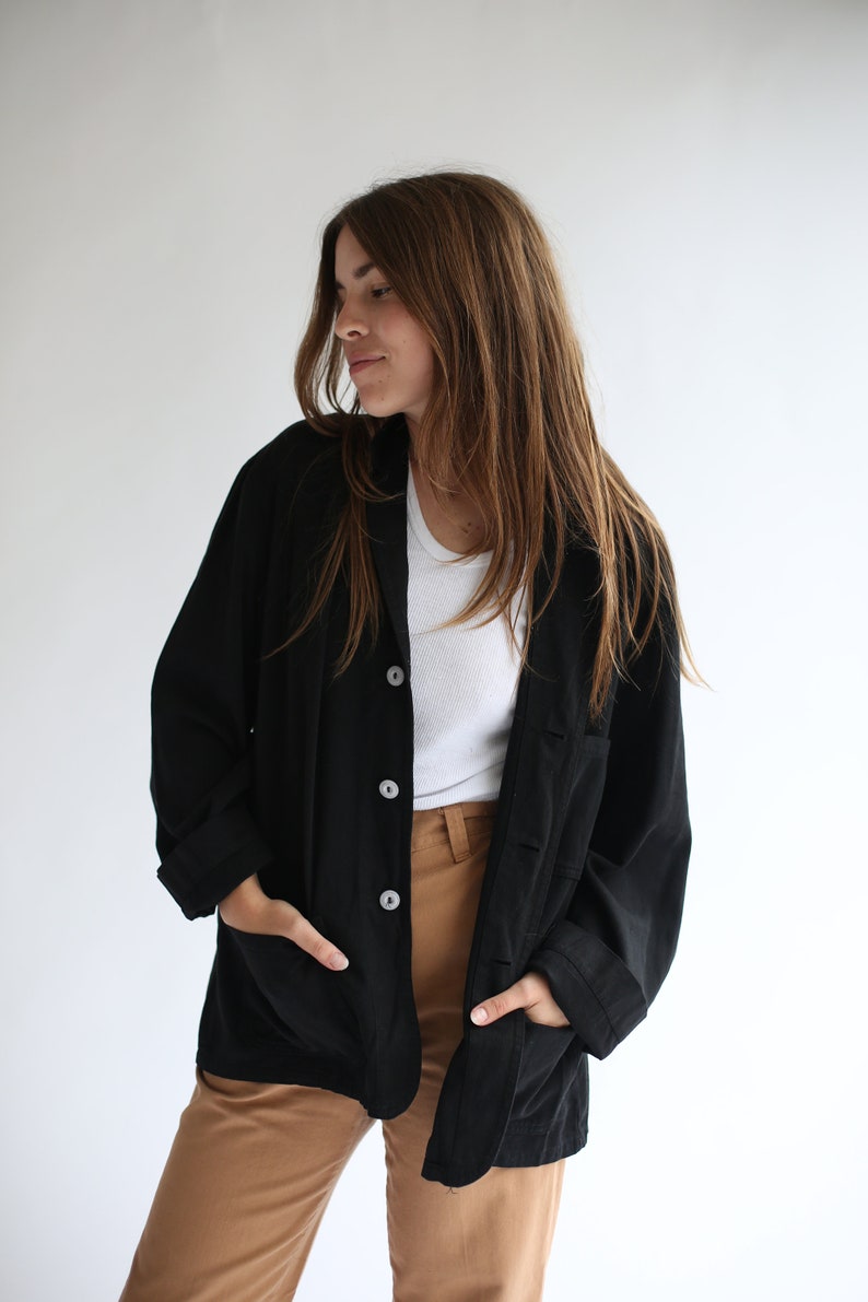 May include: A black long-sleeved button-up shirt with a relaxed fit and pockets. The shirt is worn over a white t-shirt and brown trousers.