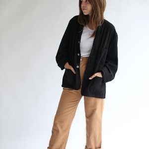 May include: A woman wearing a black button-up jacket over a white t-shirt, brown trousers, and black ankle boots.