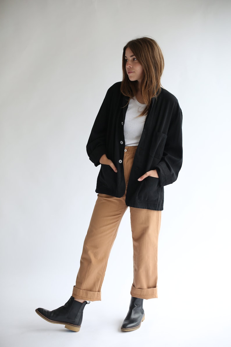 May include: A woman wearing a black button-up jacket over a white t-shirt, brown trousers, and black ankle boots.