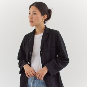 Op de afbeelding: Een vrouw draagt een zwarte utility-jas over een wit t-shirt en een blauwe jeans.
