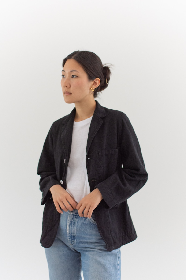 Op de afbeelding: Een vrouw draagt een zwarte utility-jas over een wit t-shirt en een blauwe jeans.