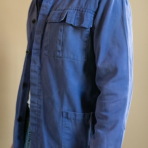 May include: A long-sleeved blue work jacket featuring a classic collar, a button-down front, and two large patch pockets on the lower front. The garment also includes a single chest pocket with a flap closure and visible stitching details.