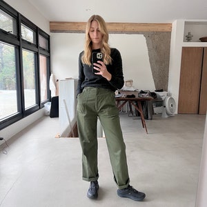 May include: A person wearing a dark grey jumper, olive green trousers, and black trainers. The trousers have a high waist and a relaxed fit. The trainers are dark and have a thick sole. The person is holding a phone.