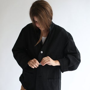 May include: A black linen jacket with a button fastening and two pockets. The jacket is undone and the person wearing it is adjusting the button.