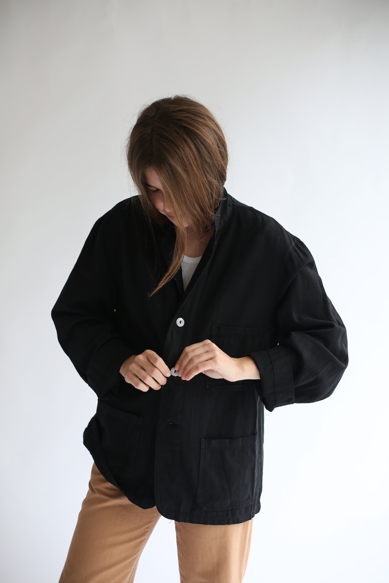 May include: A black linen jacket with a button fastening and two pockets. The jacket is undone and the person wearing it is adjusting the button.
