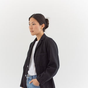 Op de afbeelding: Een vrouw draagt een zwart blazer over een wit t-shirt en een blauwe jeans. Ze staat voor een witte achtergrond.