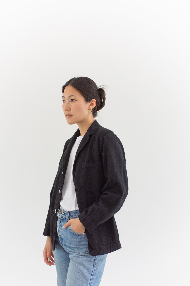 Op de afbeelding: Een vrouw draagt een zwart blazer over een wit t-shirt en een blauwe jeans. Ze staat voor een witte achtergrond.