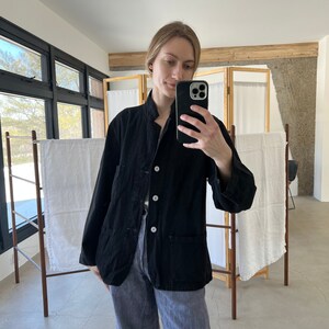 May include: A black cotton jacket with a mandarin collar, button closure, and two patch pockets. The jacket is worn over a pair of blue jeans.