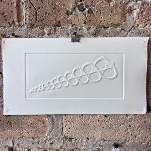 May include: A white rectangular artwork with a raised, repeating spiral design. The artwork is attached to a brick wall with a metal clip. The design starts small and expands, creating a visual flow. The paper has a textured surface.