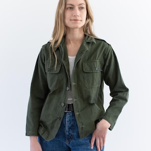 Può includere: Giacca verde oliva in stile militare con due tasche sul petto e chiusure con bottoni. La giacca è indossata sopra una canotta bianca e abbinata a jeans blu. È visibile una cintura nera.