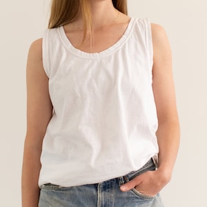 May include: A white sleeveless tank top with a scoop neck and a relaxed fit. The tank top is made of a soft, lightweight fabric and has a slightly distressed look.
