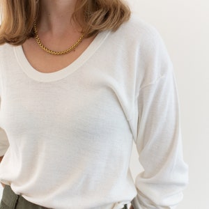 May include: A long-sleeve white crew neck shirt with a soft texture, paired with olive green trousers featuring a button closure at the waist. A gold chain necklace and small gold hoop earrings are worn with the shirt, which has a relaxed fit.