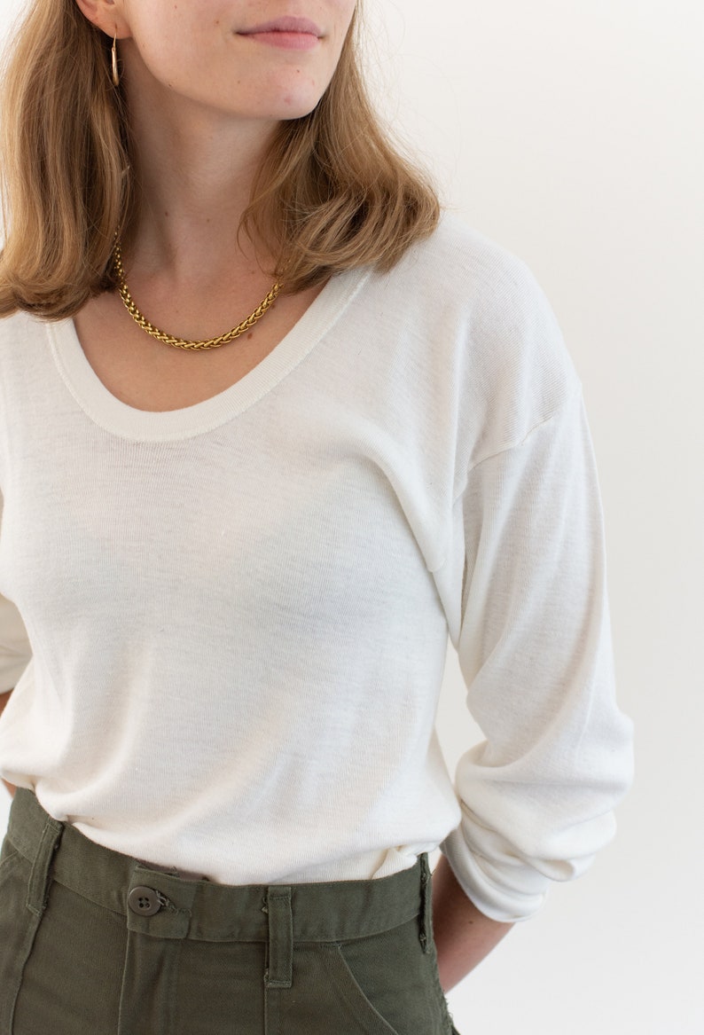 May include: A long-sleeve white crew neck shirt with a soft texture, paired with olive green trousers featuring a button closure at the waist. A gold chain necklace and small gold hoop earrings are worn with the shirt, which has a relaxed fit.