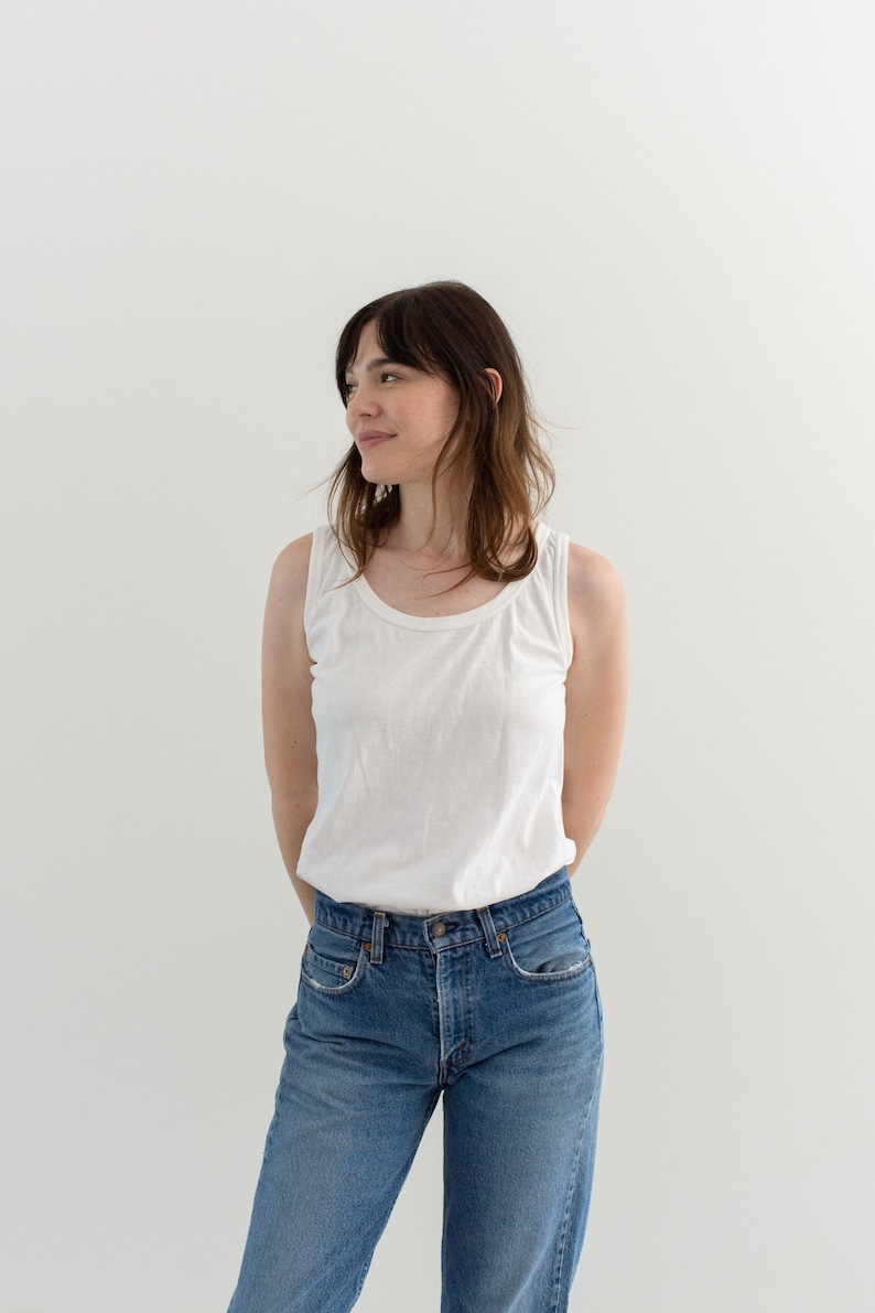 May include: A woman wearing a white tank top and blue jeans. She is standing in front of a white wall and looking to the side.