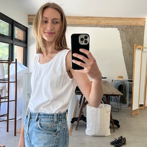 Puede incluir: Una mujer con un top blanco sin mangas y pantalones vaqueros azules claros. Est&aacute; tom&aacute;ndose una selfie con un tel&eacute;fono inteligente negro.