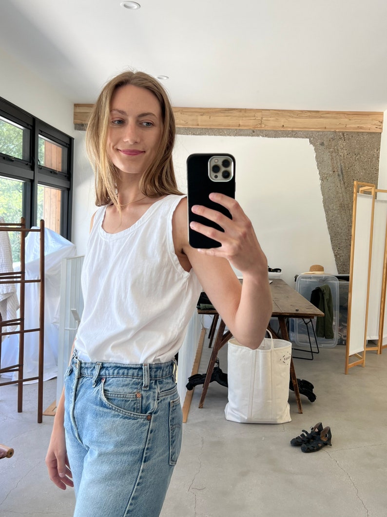 Puede incluir: Una mujer con un top blanco sin mangas y pantalones vaqueros azules claros. Est&aacute; tom&aacute;ndose una selfie con un tel&eacute;fono inteligente negro.