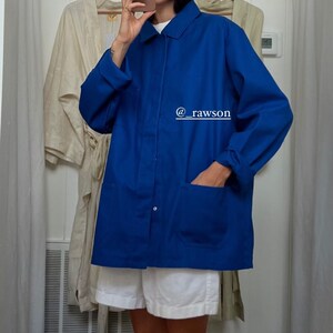May include: A woman wearing a white shirt and white shorts is standing in a dressing room. She is wearing a blue utility jacket with a single button closure and two large pockets. The jacket has a rolled up cuff. The text "@rawson" is written on the jacket.