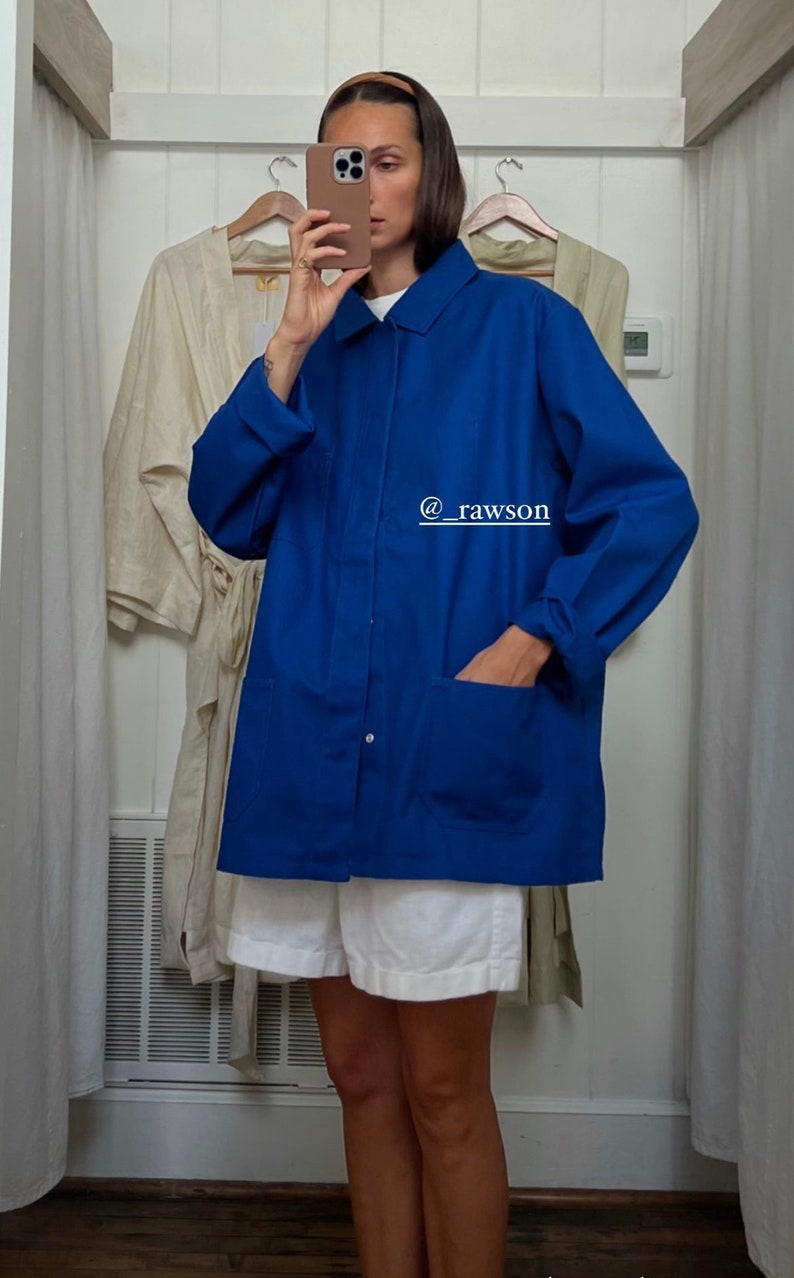 May include: A woman wearing a white shirt and white shorts is standing in a dressing room. She is wearing a blue utility jacket with a single button closure and two large pockets. The jacket has a rolled up cuff. The text "@rawson" is written on the jacket.