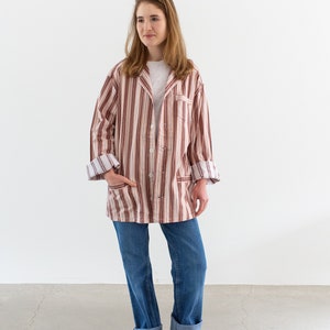 May include: A person wearing a striped, button-down jacket in shades of brown, red, and white, paired with blue jeans and gray boots. The jacket has a relaxed fit and rolled-up sleeves. The outfit is casual and comfortable.