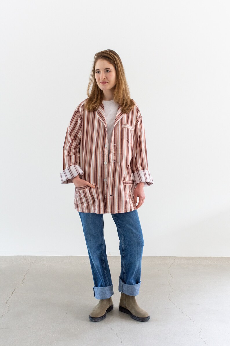 May include: A person wearing a striped, button-down jacket in shades of brown, red, and white, paired with blue jeans and gray boots. The jacket has a relaxed fit and rolled-up sleeves. The outfit is casual and comfortable.