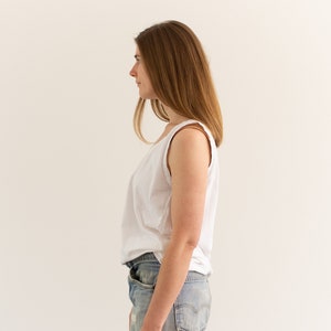 May include: A woman wearing a white tank top and light blue jeans stands facing a white wall. Her hair is long and brown.