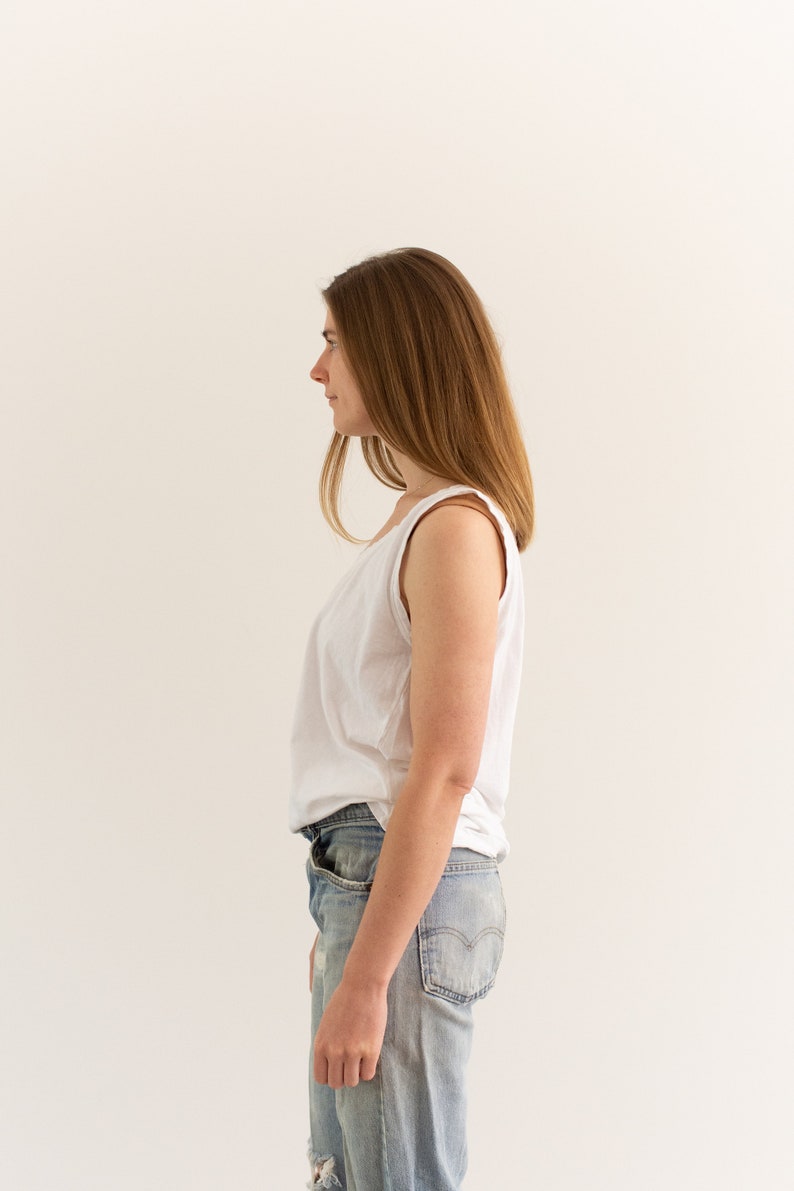 May include: A woman wearing a white tank top and light blue jeans stands facing a white wall. Her hair is long and brown.