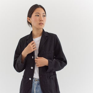 Op de afbeelding: Een vrouw draagt een zwart utility-jack met witte knopen en een wit T-shirt eronder. Ze draagt een blauwe jeans en heeft haar haar naar achteren getrokken.