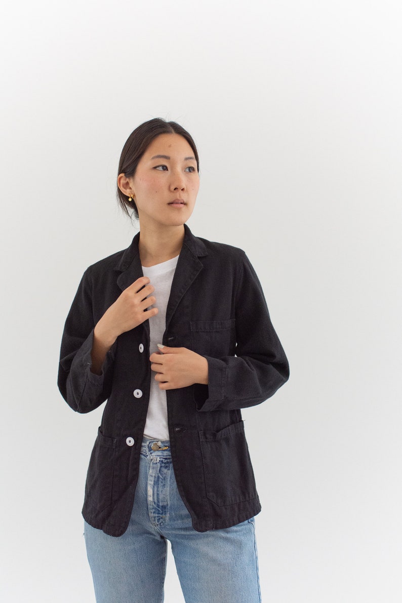 Op de afbeelding: Een vrouw draagt een zwart utility-jack met witte knopen en een wit T-shirt eronder. Ze draagt een blauwe jeans en heeft haar haar naar achteren getrokken.
