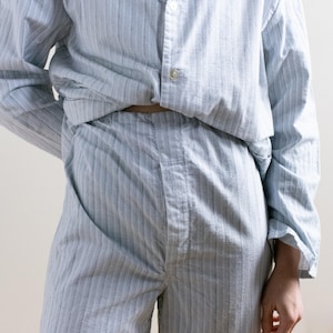 May include: Light blue and white striped pajama set. The long-sleeved shirt has a button-down front and a collar. The matching pants have a relaxed fit. The fabric has a subtle herringbone pattern. The pajama set is made of a soft, lightweight material.