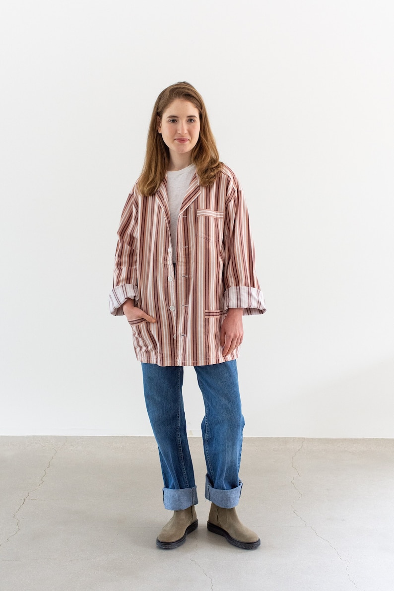 May include: A button-down, striped shirt in shades of red, brown, and cream, paired with blue jeans. The shirt has a relaxed fit and is worn over a white t-shirt. The jeans are cuffed at the ankles, and the person is wearing gray boots.