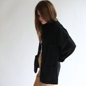 May include: A woman wearing a black long-sleeved shirt with a collar and brown trousers. The shirt has a relaxed fit and a slightly oversized look.