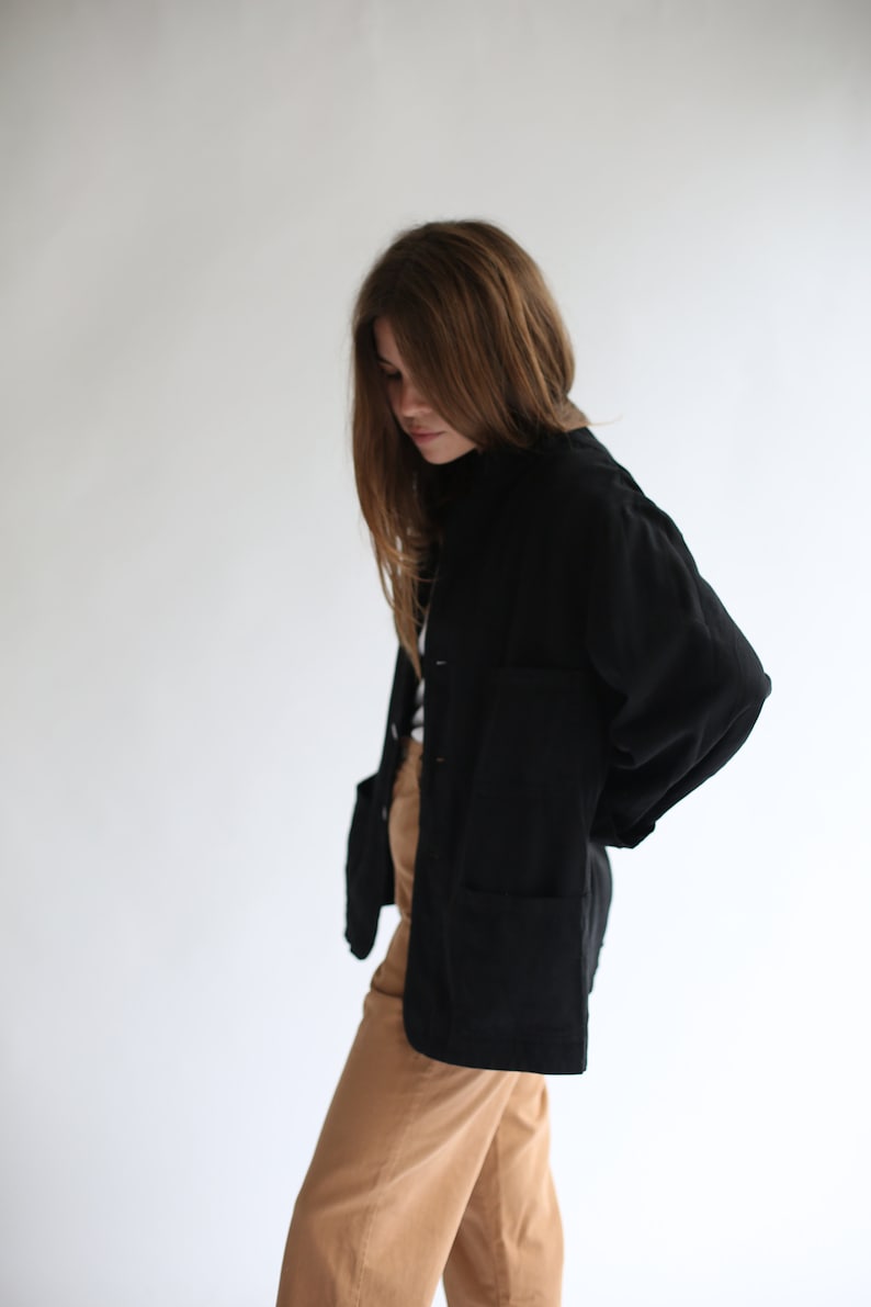 May include: A woman wearing a black long-sleeved shirt with a collar and brown trousers. The shirt has a relaxed fit and a slightly oversized look.