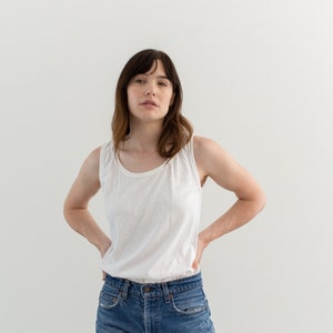 May include: A woman wearing a white tank top and blue jeans. She is standing in front of a white wall.