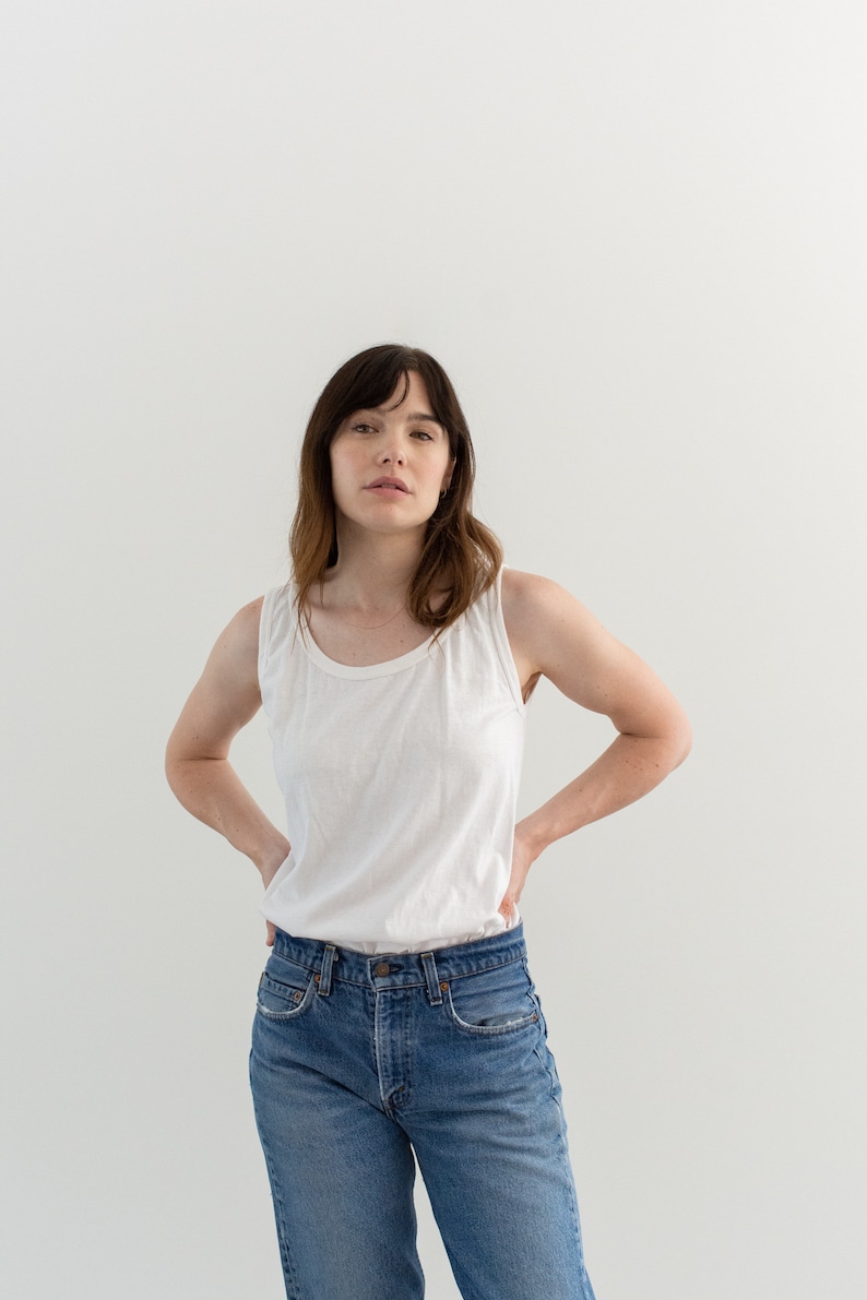 May include: A woman wearing a white tank top and blue jeans. She is standing in front of a white wall.