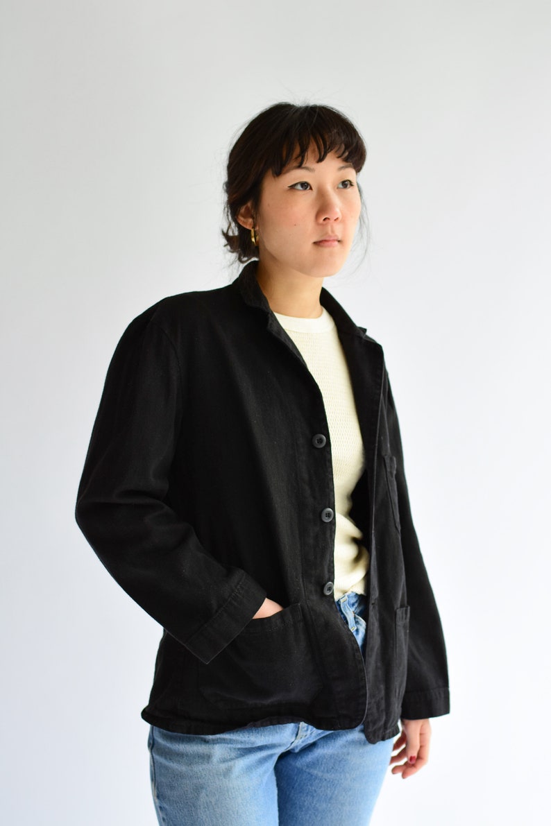 May include: A black utility jacket with a button closure and a single pocket on the left side. The jacket is worn over a white shirt and blue jeans.