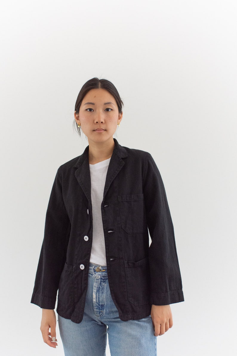 Op de afbeelding: Een zwart utility-jack met een wit overhemd met knopen eronder en een blauwe jeans. Het jack heeft twee borstzakken en twee lagere zakken.