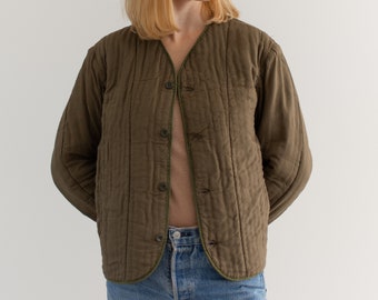 Puede incluir: Una chaqueta acolchada de color verde oliva oscuro con cierre de botones y cuello en V. La chaqueta tiene un tejido ligeramente texturizado y un ribete verde alrededor de los bordes.