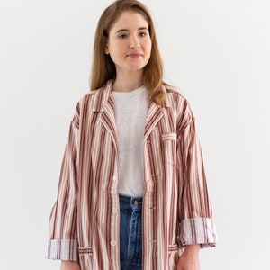 May include: A striped button-down shirt in shades of brown, beige, and white. The shirt has a classic collar, front pockets, and is worn over a white t-shirt and blue jeans. The sleeves are rolled up.