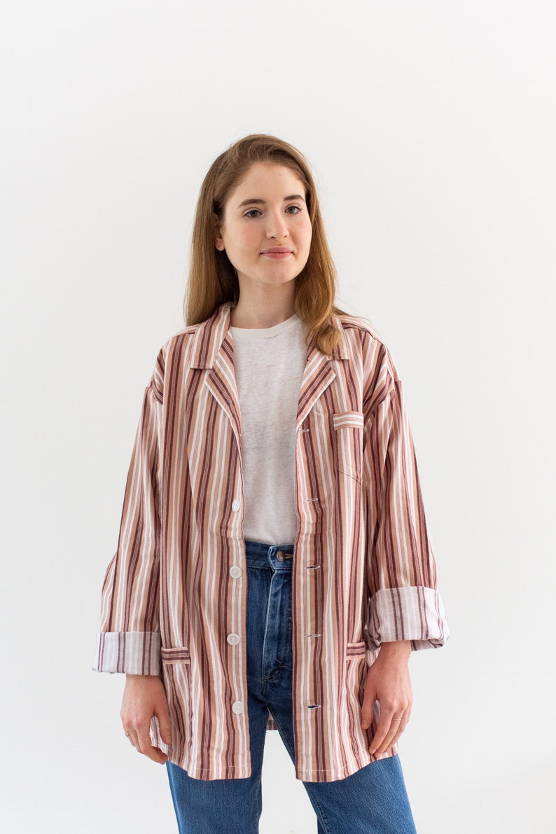 May include: A striped button-down shirt in shades of brown, beige, and white. The shirt has a classic collar, front pockets, and is worn over a white t-shirt and blue jeans. The sleeves are rolled up.