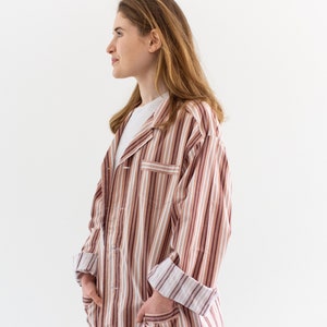 May include: A striped button-down shirt in shades of brown, white, and burgundy. The shirt has a collared neckline, long sleeves, and two front pockets. The sleeves are cuffed, and the shirt is worn over a white t-shirt and blue jeans.