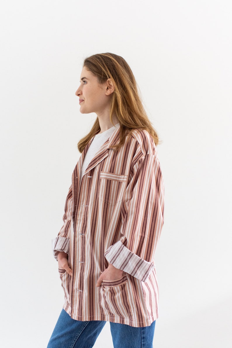 May include: A striped button-down shirt in shades of brown, white, and burgundy. The shirt has a collared neckline, long sleeves, and two front pockets. The sleeves are cuffed, and the shirt is worn over a white t-shirt and blue jeans.
