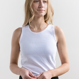 Peut inclure: Un débardeur blanc à col rond. Le haut sans manches est fait d'un tissu doux et clair. Le modèle porte un pantalon noir. Le fond est blanc uni.