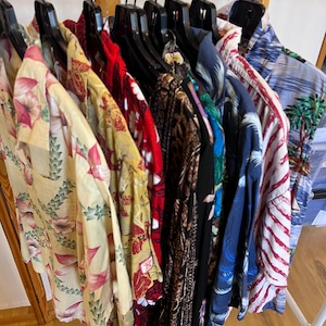 May include: A collection of colorful button-down shirts hanging on a rack. The shirts feature various tropical and floral patterns in shades of red, yellow, blue, and green. The shirts are made of a lightweight fabric.