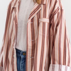 May include: A button-down striped shirt in shades of brown, white, and tan. The shirt has a chest pocket and rolled-up sleeves. The model is wearing a white t-shirt and blue jeans.