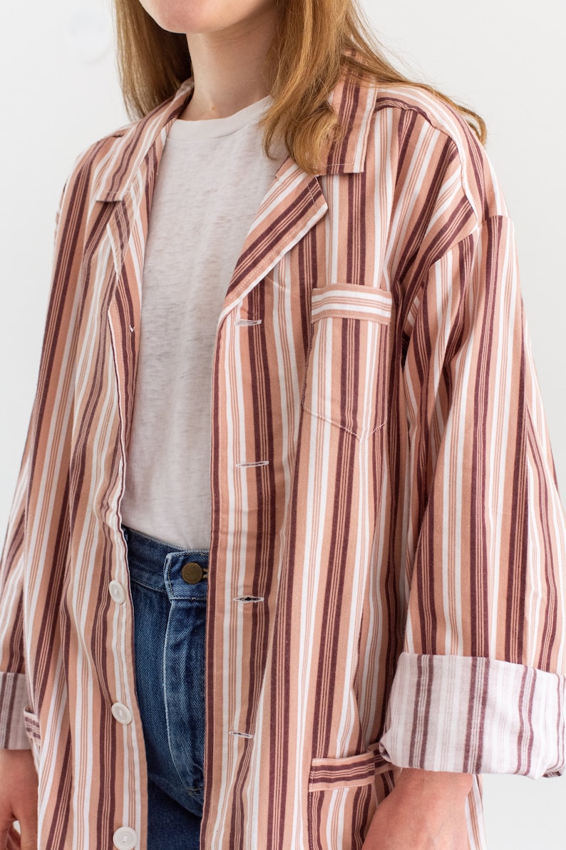 May include: A button-down striped shirt in shades of brown, white, and tan. The shirt has a chest pocket and rolled-up sleeves. The model is wearing a white t-shirt and blue jeans.