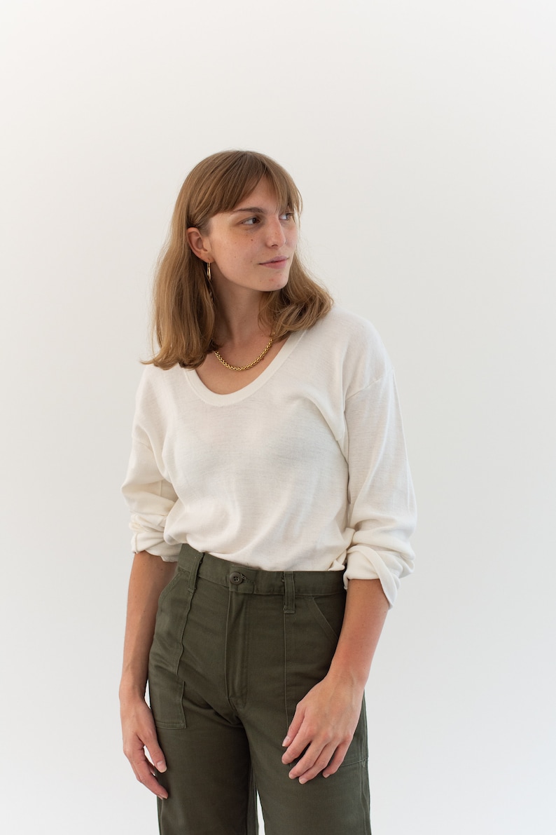 May include: A long-sleeved, off-white scoop neck shirt paired with olive green utility pants featuring a button closure and front patch pockets. The shirt has a relaxed fit with sleeves that are rolled up at the forearms, and the pants have a high-waisted design.