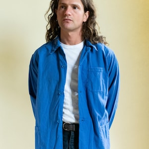 May include: A bright blue long-sleeve button-down shirt featuring a classic collar, a single chest pocket, and two lower front pockets. The shirt is worn over a plain white t-shirt and paired with dark blue denim jeans secured by a black leather belt.